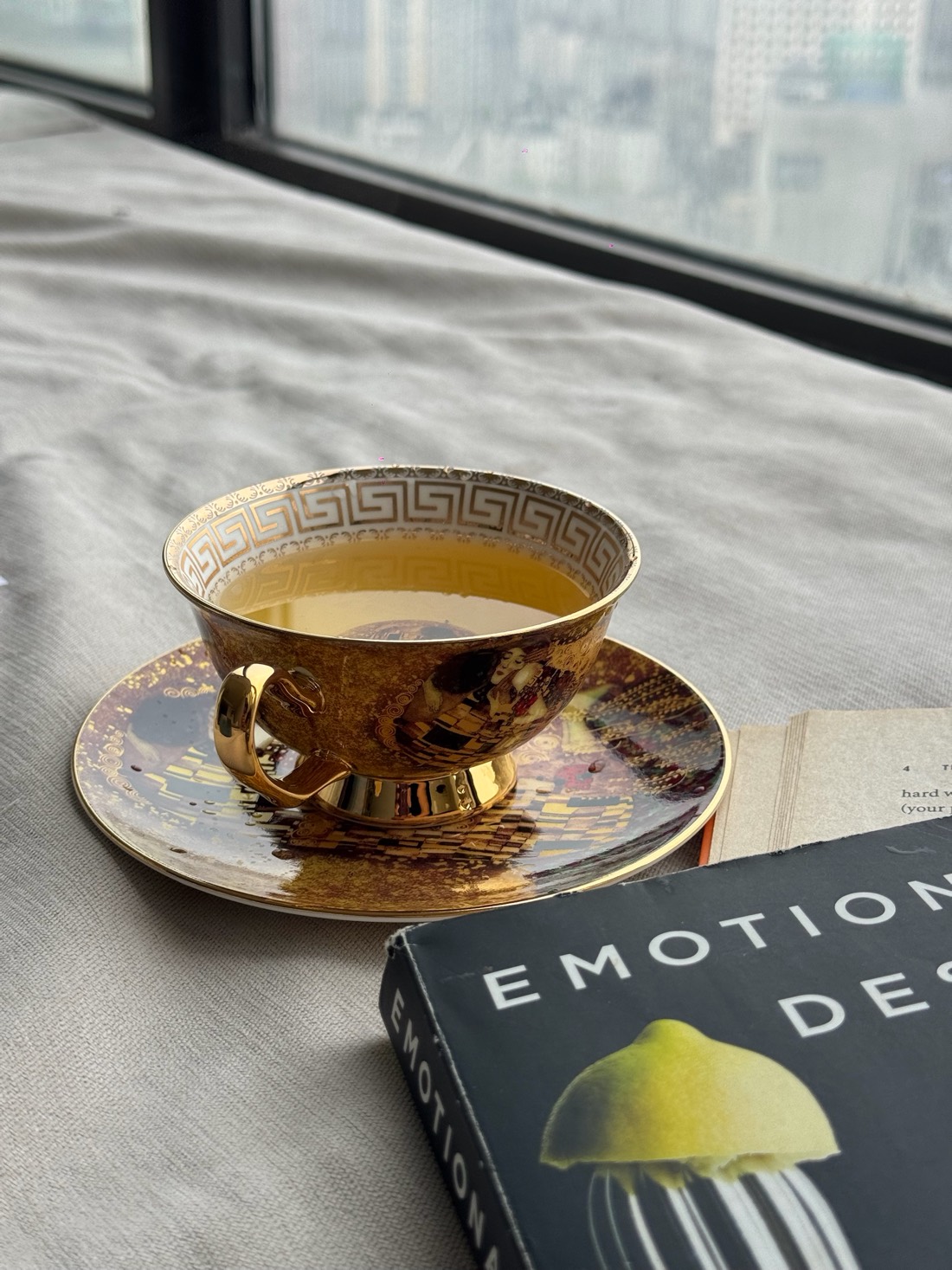 A gold bone china teacup with a Greek key border inside the rim, on a matching saucer, beside a window and a copy of Emotional Design.